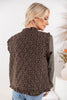 A woman shown from the back wearing a sleeveless quilted vest with a brown floral pattern and ruffled trim, layered over a dark olive green long-sleeve top and blue jeans.