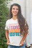 A smiling woman with long wavy brown hair wearing a white graphic t-shirt that reads 'PUMPKIN S'MORE HAYRIDE BONFIRES' in stacked colorful letters, paired with blue jeans, standing outdoors in front of a white brick wall and green foliage.