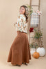 A woman modeling a long-sleeve cream floral shirt and a brown corduroy maxi skirt, standing in a studio decorated with a ladder, pumpkins, and flowers.