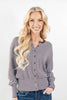 A smiling blonde woman wearing a long-sleeve button-front top with thin brown and light blue horizontal stripes, paired with blue jeans, against a plain white studio background.