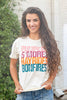 A smiling woman with long wavy brown hair stands outdoors wearing a cream-colored graphic t-shirt with the words 'Pumpkin S'mores Hayrides Bonfires' printed in pink, purple, orange, and blue. She is also wearing blue jeans.