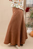 A wom wearing a long, flowing brown skirt paired with a white floral top, standing in a styled indoor setting with plants and vases in the background.n