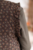 Close-up of a dark brown quilted vest with a small orange and green floral pattern, featuring a ruffled trim, worn over a green top.