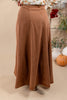 A woman wearing a long, flowing brown skirt with a floral long-sleeve top, standing against a warm beige background with decorative pumpkins and flowers.