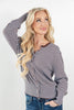 A blonde woman wearing a brown and light blue striped long-sleeve button-up top with a split neckline, paired with blue jeans, posing against a light gray studio background.