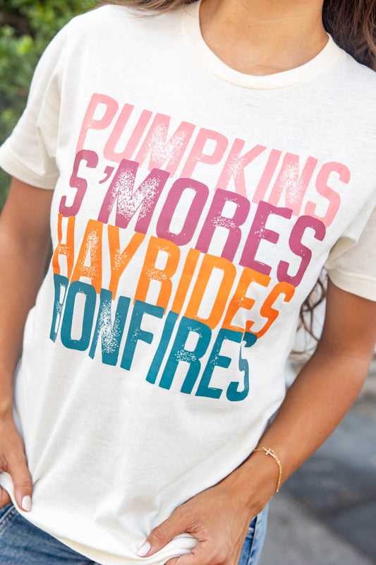 Close up shot of a woman wearing a cream-colored graphic t-shirt with the words 'Pumpkin S'mores Hayrides Bonfires' printed in pink, purple, orange, and blue. 