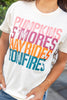 Close up shot of a woman wearing a cream-colored graphic t-shirt with the words 'Pumpkin S'mores Hayrides Bonfires' printed in pink, purple, orange, and blue. 