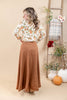 A woman stands back wearing a long-sleeve white floral blouse and a high-waisted, ankle-length brown skirt. She is posed in front of a beige studio backdrop decorated with a wooden ladder, potted pumpkins, and floral arrangements.