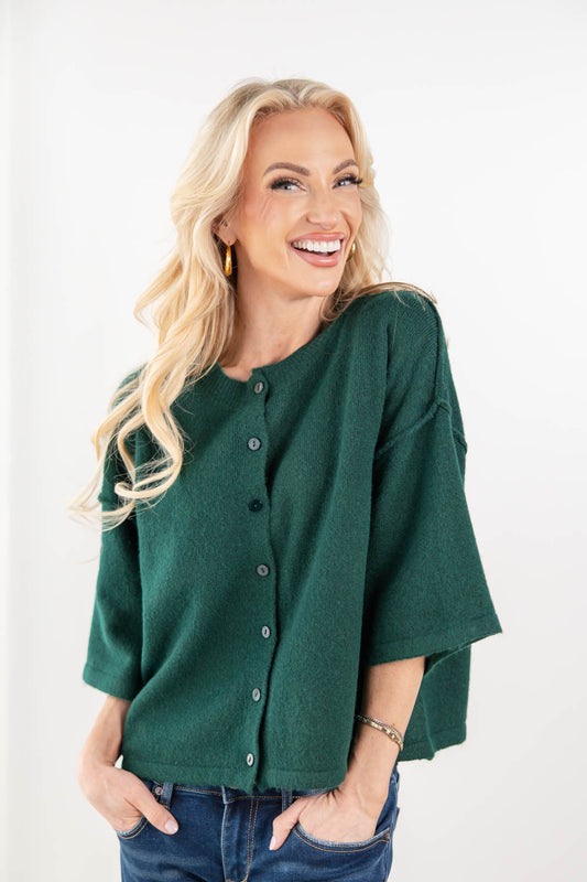A smiling blonde woman wearing a dark green button-front knit top with wide three-quarter sleeves and blue jeans, standing against a plain white studio background.