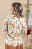 A woman shown from the back wearing a cream knit sweater with an orange, yellow, and green floral pattern, paired with brown pants in a warm-toned indoor setting.
