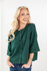 A smiling blonde woman wearing a dark green button-front knit top with wide three-quarter sleeves and blue jeans, standing against a plain white studio background.