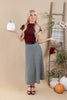 A woman stands in a studio setting wearing a burgundy short-sleeve mock-neck top and a black and white gingham midi skirt. She holds a white decorative pumpkin in one hand and gestures with the other.