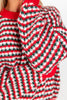 A close-up of a person wearing a chunky knit sweater with a festive red, green, and white patterned design and solid red ribbed collar and cuffs.