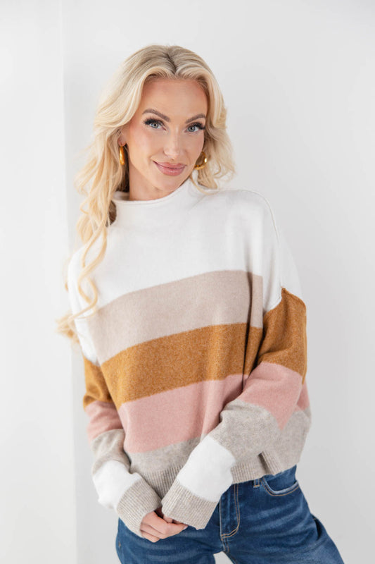 A woman with blonde hair wearing a color-blocked knit sweater with horizontal stripes of white, beige, mustard yellow, pink, and light gray, paired with blue jeans.