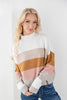 A woman with blonde hair wearing a color-blocked knit sweater with horizontal stripes of white, beige, mustard yellow, pink, and light gray, paired with blue jeans.