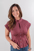 A smiling woman with brown hair wearing a cap-sleeve, mock-neck, keyhole button-front blouse in a mauve color, paired with blue jeans, against a plain light gray studio background.