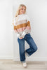 A woman posing in a white, beige, and brown striped knit sweater with wide-leg blue jeans and white sneakers, standing against a white wall.