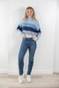 A woman modeling a light blue, navy, and gray striped mock-neck sweater, paired with blue jeans and white sneakers, standing against a white wall.