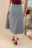 A woman wearing a black and white gingham midi skirt with a dark waistband, paired with a burgundy top and burgundy block-heel sandals, standing against a beige studio backdrop with decorative autumn props.