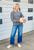 A woman stands outdoors in front of a white garage door wearing a gray long-sleeve crewneck sweatshirt with a small white ghost graphic on the chest, blue wide-leg jeans, and white slip-on shoes. She is holding a small carved jack-o'-lantern.