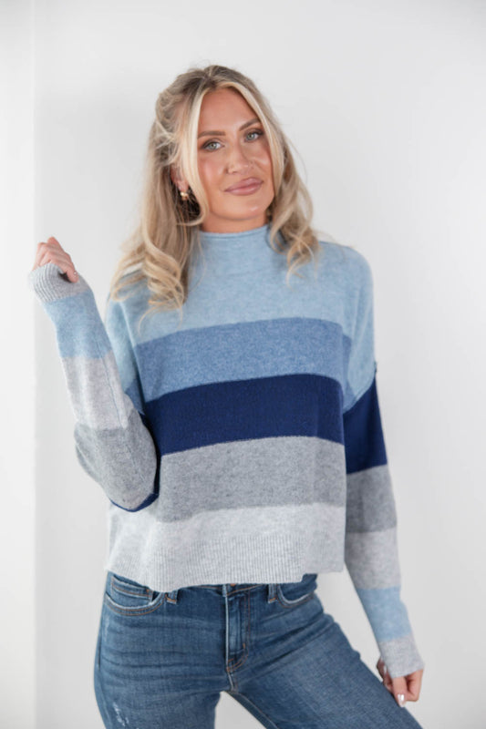 A woman wearing a color-blocked knit sweater with horizontal stripes in light blue, navy, and heather gray, paired with blue jeans, against a plain white studio background.