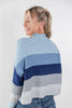 A woman wearing a color-block knit sweater with horizontal stripes in light blue, navy, and gray, shown from a three-quarters back view against a plain white studio background.