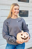 A smiling blonde woman wearing a gray crewneck sweatshirt with 'Spooky Season' printed on the chest and bat motifs on the sleeve, holding a carved jack-o'-lantern pumpkin in front of a white siding background.