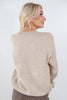 A woman with blonde hair is shown from the back wearing a light beige knit crewneck sweater and brown pants against a plain white background.