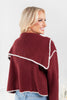 A woman wearing a burgundy ribbed knit cropped sweater with a wide triangular collar trimmed in white picot trim, shown from a three-quarters back view.