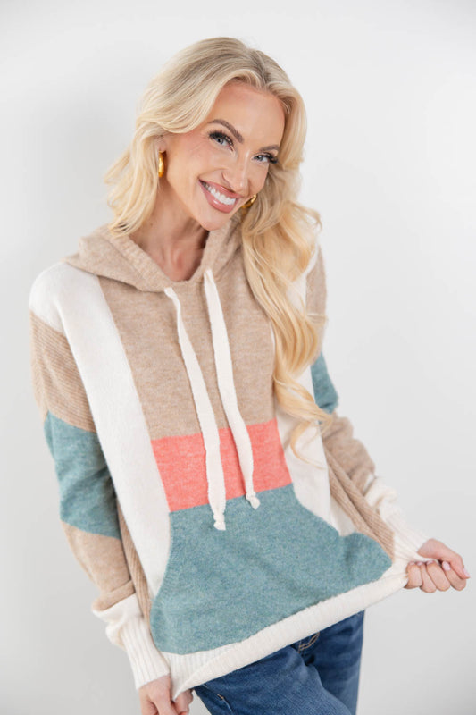 A smiling blonde woman models a color-block knit hoodie with beige, white, teal, and coral pink sections, paired with blue jeans against a plain light gray studio background.