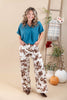 A woman modeling a teal short-sleeve blouse and brown-and-white cow-print wide-leg pants, standing in a studio with a beige backdrop, a wooden ladder, and fall decor including pumpkins and flowers.