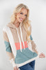A smiling blonde woman models a color-block knit hoodie with beige, white, teal, and coral pink sections, paired with blue jeans against a plain light gray studio background.
