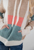 A close-up of a woman wearing a color-block knit hoodie with cream, tan, coral pink, and teal sections, with the front pockets and white drawstrings visible.