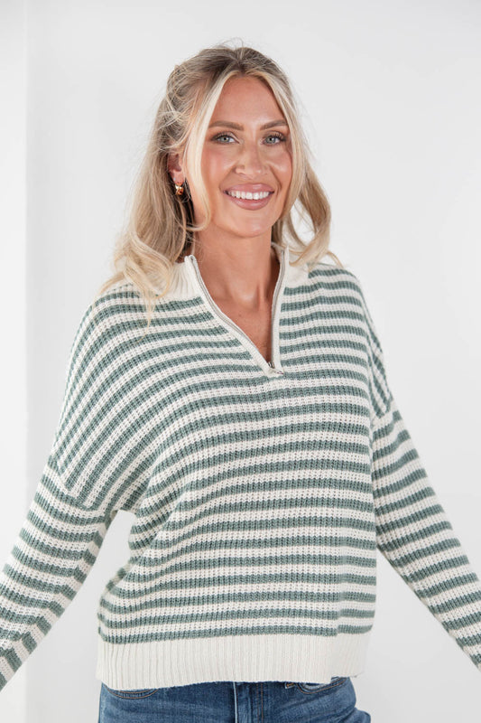 A smiling blonde woman wearing a green and off-white striped knit sweater with a half-zip neckline, paired with blue jeans, against a plain white background.