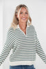 A smiling blonde woman wearing a green and off-white striped knit sweater with a half-zip neckline, paired with blue jeans, against a plain white background.