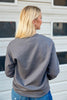 A woman with blonde hair is shown from behind wearing a dark gray crewneck sweatshirt and blue jeans, standing in front of a white siding wall.