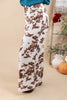A person wearing wide-leg pants with a brown and white cow print pattern, paired with a teal top. The photo is cropped from the waist down, set against a warm beige background with decorative props like a small pumpkin and white vases.