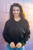 A smiling young woman with long brown hair stands against a purple-painted brick wall, wearing a black long-sleeve henley-style knit top and blue jeans.