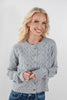 A smiling blonde woman wearing a light gray pointelle-knit cardigan with brown buttons and blue jeans, photographed against a plain light gray studio background.