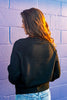A woman with long brown hair is shown from behind wearing a black knit sweater and light blue jeans, standing against a purple-painted brick wall with warm sunlight highlighting her hair.