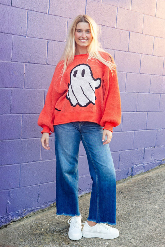A woman stands against a purple painted brick wall wearing a bright orange crewneck sweater with a large white ghost graphic, blue wide-leg jeans, and white sneakers.