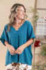 A woman wearing a teal blue short-sleeve V-neck top, standing in a warm-toned indoor setting with a wooden ladder and home decor in the background.