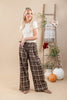 A woman stands in a studio wearing brown and beige plaid wide-leg pants with a white t-shirt. The background features a beige backdrop, a wooden ladder decorated with a plaid throw blanket and flowers, and two pumpkins on the floor.