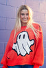 A woman stands against a purple painted brick wall wearing a bright orange crewneck sweater with a large white ghost graphic and blue jeans.