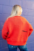 A woman with blonde hair is shown from behind, wearing a bright orange knit sweater and blue jeans, standing against a purple painted brick wall.