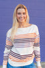A smiling blonde woman wearing a cream-colored knit sweater with horizontal stripes in orange and purple, standing against a purple painted brick wall.