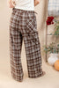A person shown from the back wearing brown and beige plaid wide-leg pants with an elastic drawstring waist, standing in a styled indoor setting with a cream wall and decorative plants.