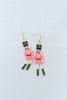 A pair of dangling earrings shaped like pink nutcracker soldiers, with gold-tone hooks, black and yellow hats, and black boots with yellow accents.