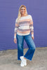 A woman stands in front of a purple brick wall wearing a cream knit sweater with orange and purple horizontal stripes, blue wide-leg jeans, and white sneakers.