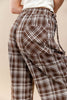 Close-up of a person wearing brown, white, and beige plaid drawstring pants with a large patch pocket, paired with a white top against a plain beige background.